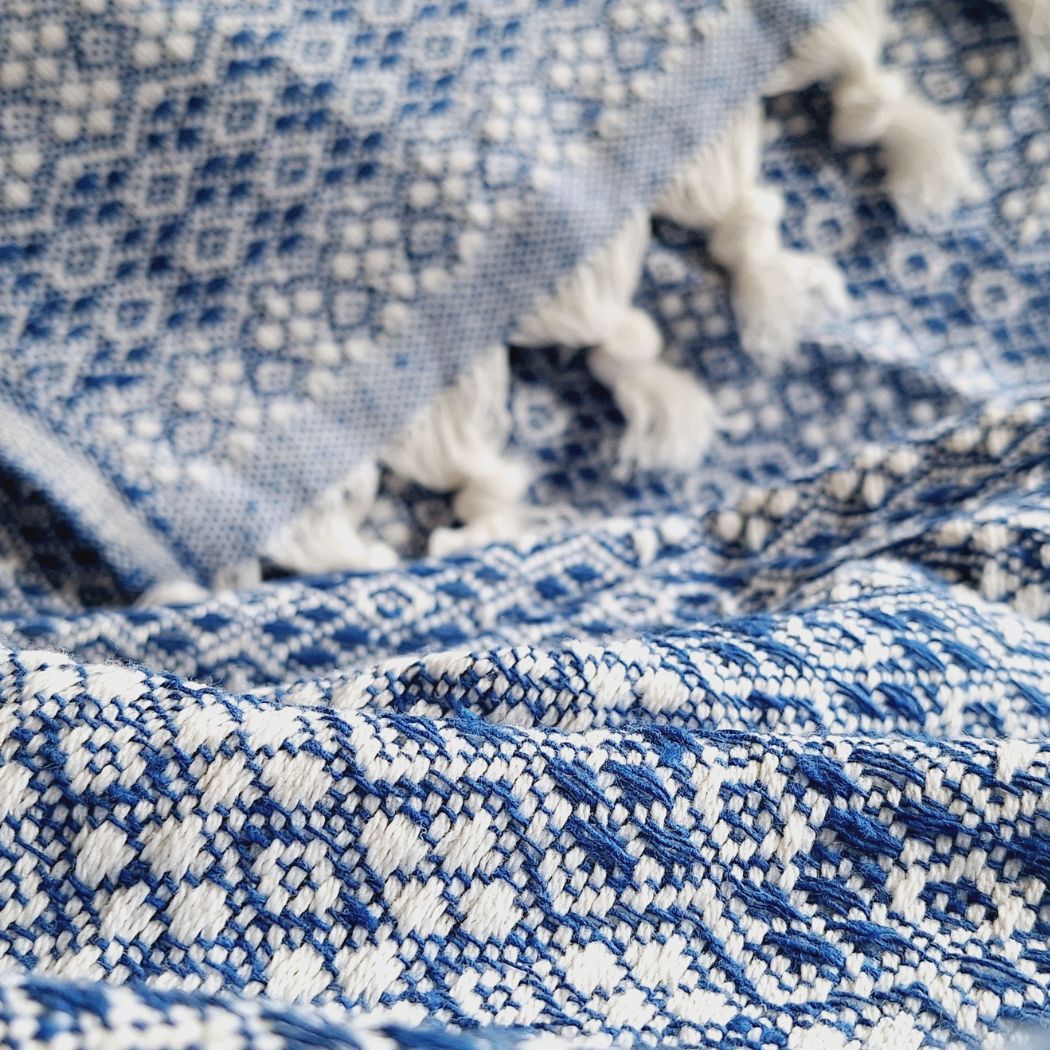 Vue rapprochée d’un couvre-lit 100% en coton, de dimensions 230 x 200 cm, avec un motif géométrique bleu et blanc. Le tissu présente une texture tissée détaillée et est agrémenté de franges nouées, apportant une touche artisanale et élégante.