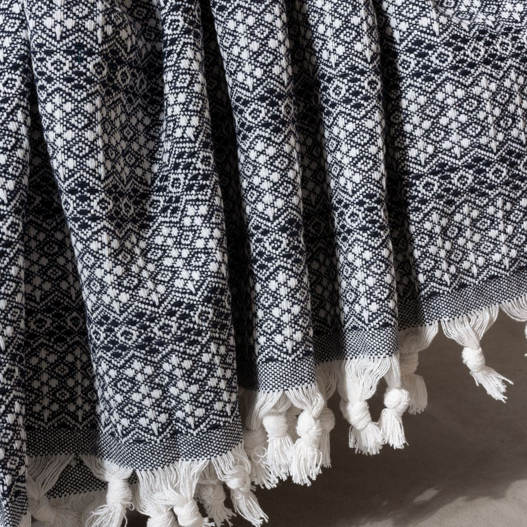 Couvre-lit 100% en coton, de dimensions 230 x 200 cm, avec un motif géométrique noir et blanc. Le tissu tissé présente une texture détaillée et est orné de franges nouées sur les bords, ajoutant une touche artisanale et élégante. Drapé avec fluidité, il met en valeur la richesse de son tissage et son design sophistiqué.