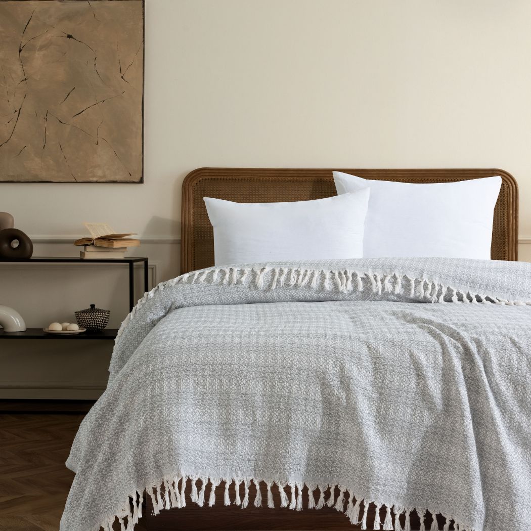 Couvre-lit 100% en coton, de dimensions 230 x 200 cm, avec un motif discret en gris et blanc. Le tissu léger et tissé avec finesse est agrémenté de franges nouées sur les bords, apportant une touche élégante et naturelle. Drapé sur un lit avec une tête en rotin et des oreillers blancs, il s’intègre parfaitement dans une décoration sobre et apaisante.