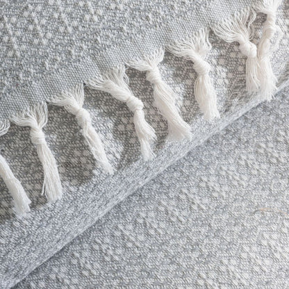 Couvre-lit 100% en coton, de dimensions 230 x 200 cm, avec un motif tissé discret en gris et blanc. Le tissu présente une texture douce et raffinée, agrémentée de franges nouées sur les bords pour une touche artisanale et élégante. Gros plan sur les détails du tissage et des finitions, mettant en valeur la qualité et le design du textile.